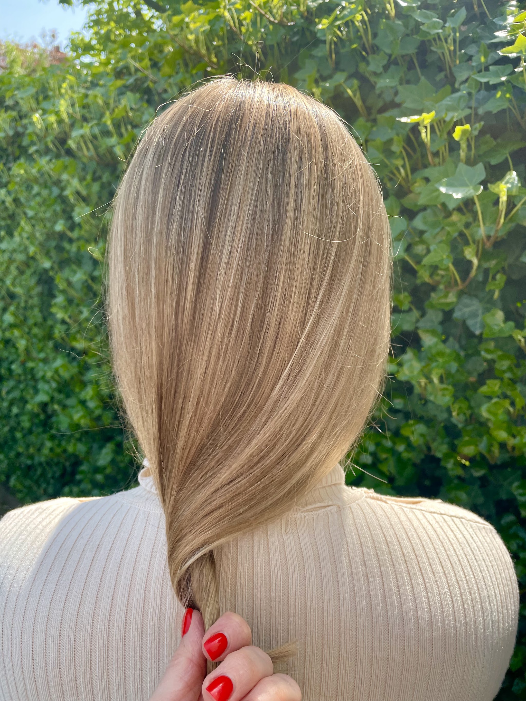 Zacht beigeblond haar buiten tegen een groene haag, rood gelakte nagels houden de punten vast